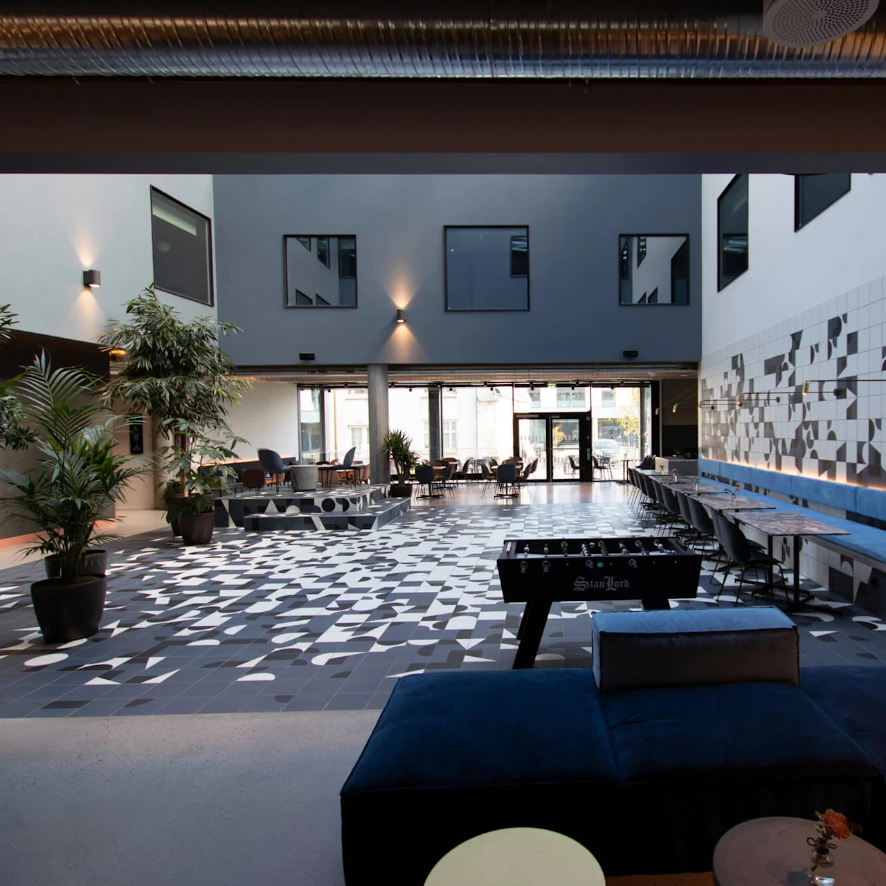 Fodboldbord står i et rummeligt, moderne atrium. Geometrisk mønstret gulv, potteplanter og siddeområder skaber en livlig atmosfære. Store vinduer oplyser rummet. Ingen tekst synlig.