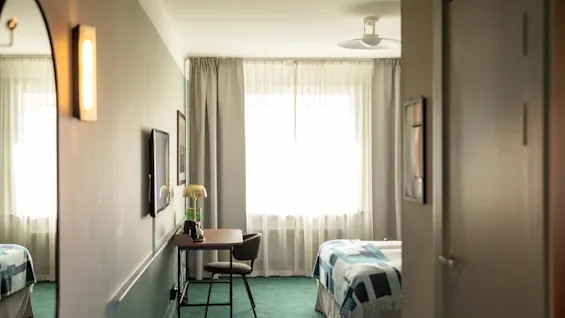 Ett prydligt arrangerat hotellrum. En säng med ett mönstrat täcke står bredvid ett skrivbord med en lampa, mot ett stort fönster med grå gardiner.