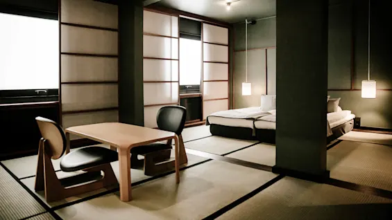 Et minimalistisk japansk stil rom med tatamigulv, et lavt bord med stoler og en seng, viser tradisjonelle og moderne elementer.