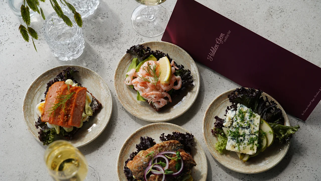 Fire tallerkener med smørbrød med forskjellige pålegg som laks, reker og urter ligger på et bord ved siden av vinglass og en rød meny med teksten "HiddenGem.se".