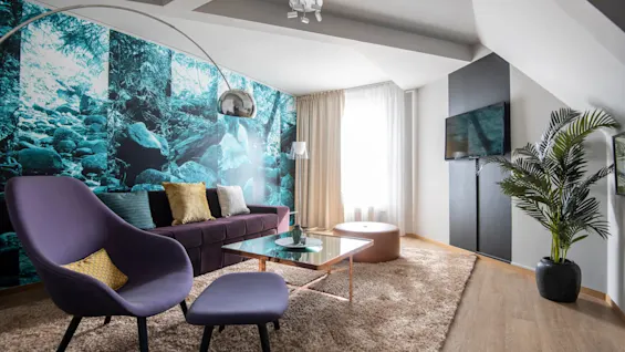 Ett modernt vardagsrum har en lila stol och matchande ottoman framför ett glas soffbord. En skogsmotivstapet pryder väggen, medan en TV är monterad bredvid en krukväxt.