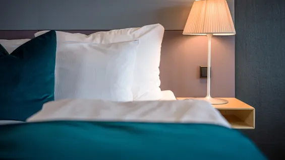 En seng med hvide og teal puder er ved siden af en tændt bordlampe i et moderne, minimalistisk soveværelsesmiljø.