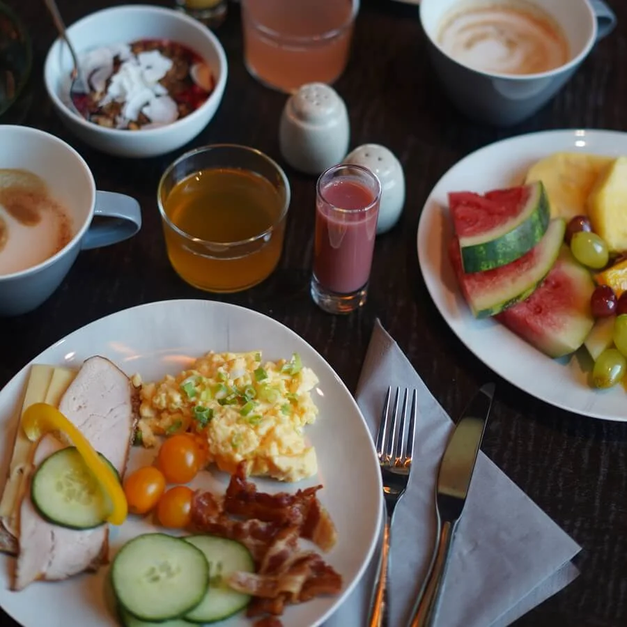 A breakfast spread on a dark table includes scrambled eggs, bacon, cheeses, fresh fruits, yogurt with coconut bits, pancakes, coffee, juices, and utensils with a napkin.