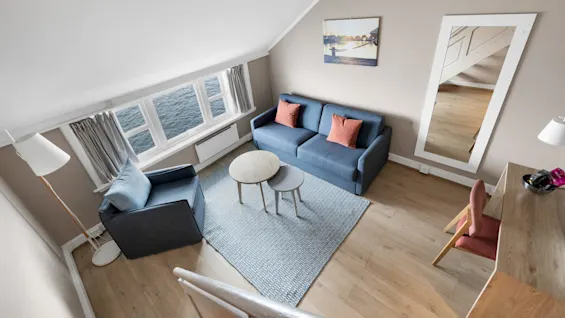 Blå sofa og stol står på et teppe mot et lite bord. Store vinduer vender mot vannet. Et speil på veggen reflekterer tregulvet. Et skrivebord med gjenstander står i nærheten.