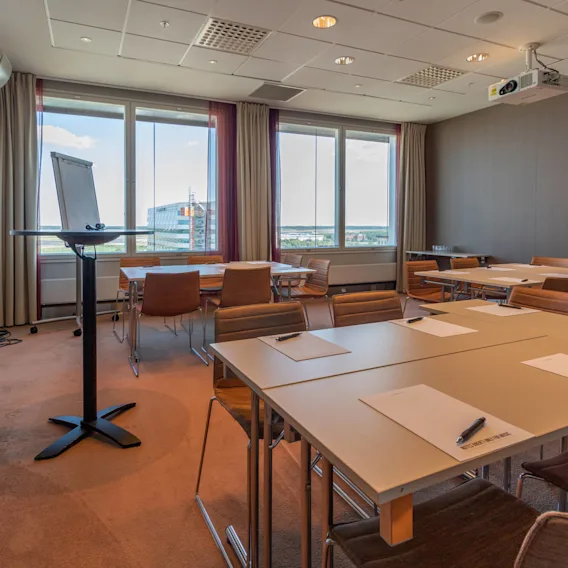 Mötesrum med rektangulära bord och bruna stolar. Anteckningsblock och pennor på borden. Golv-till-tak-fönster visar ett stadslandskap. Whiteboard och en takmonterad projektor finns. Ingen text synlig.
