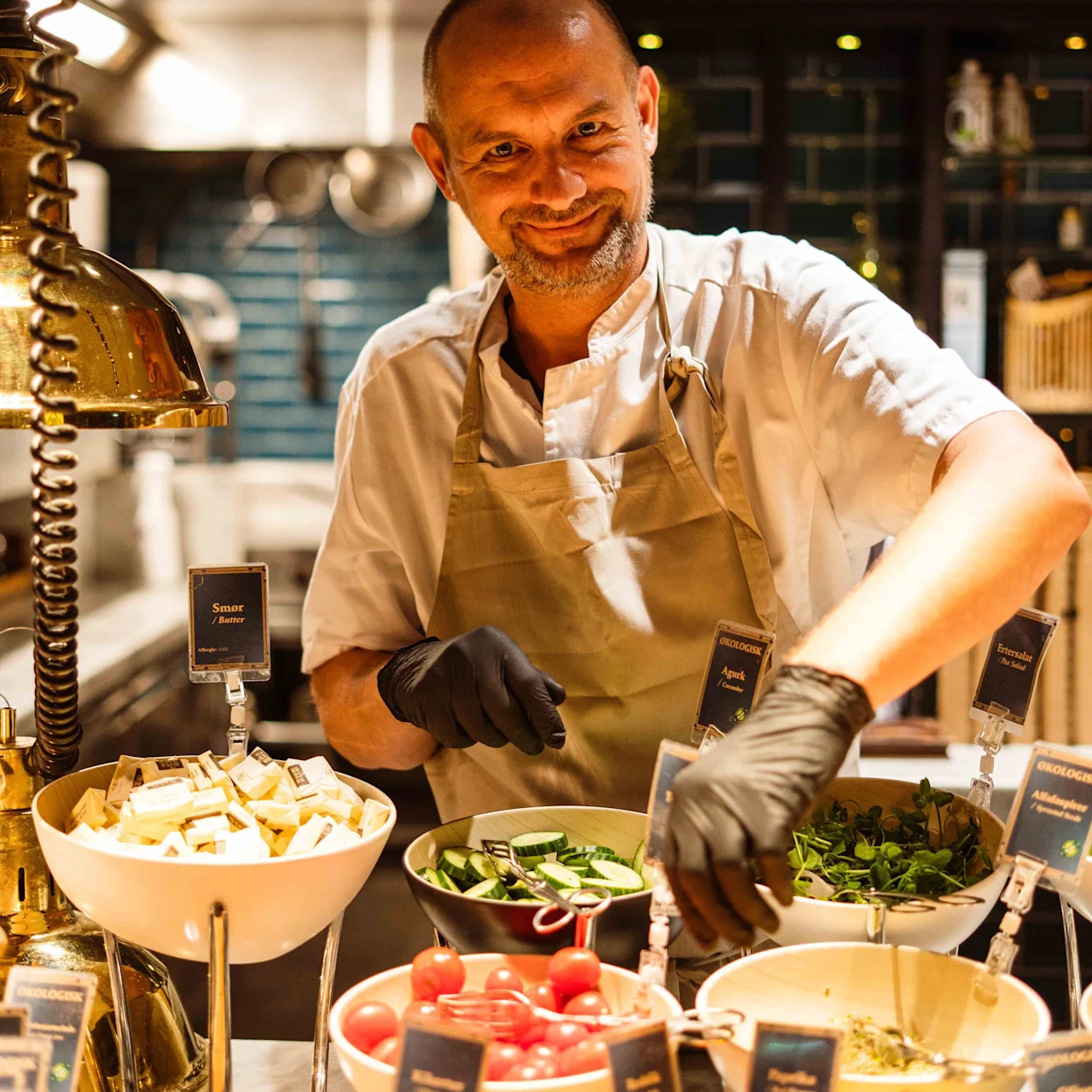 Chef prepares salad station wearing an apron and black gloves in a well-lit kitchen. Various labeled bowls contain ingredients like cherry tomatoes, cucumber slices, and herbs. Background features cooking utensils.
