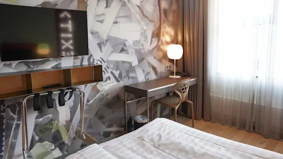 Et moderne hotellrom med flatskjerm-TV, et skrivebord med en lampe, en unik stol, gjennomsiktige gardiner og tapet med avisutklipp.