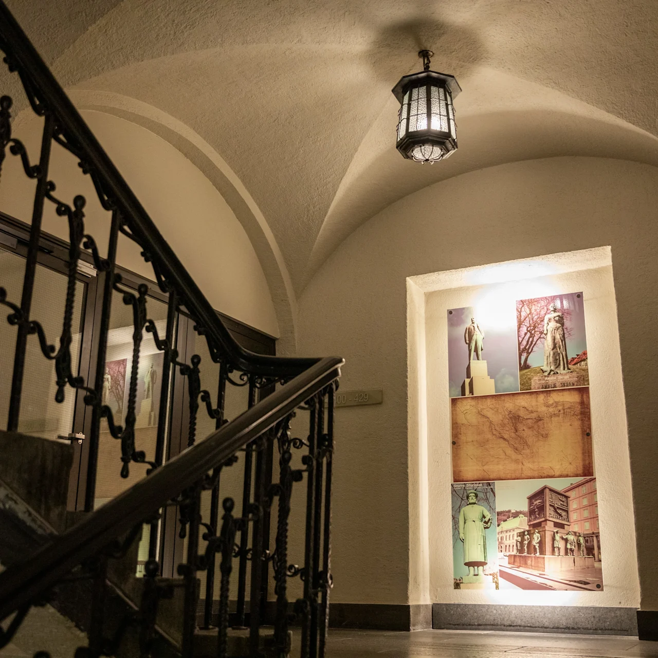 A staircase with ornate metal railings leads upward in an arched hallway. A lit display on the wall shows vintage travel posters. A hanging light fixture illuminates the area.