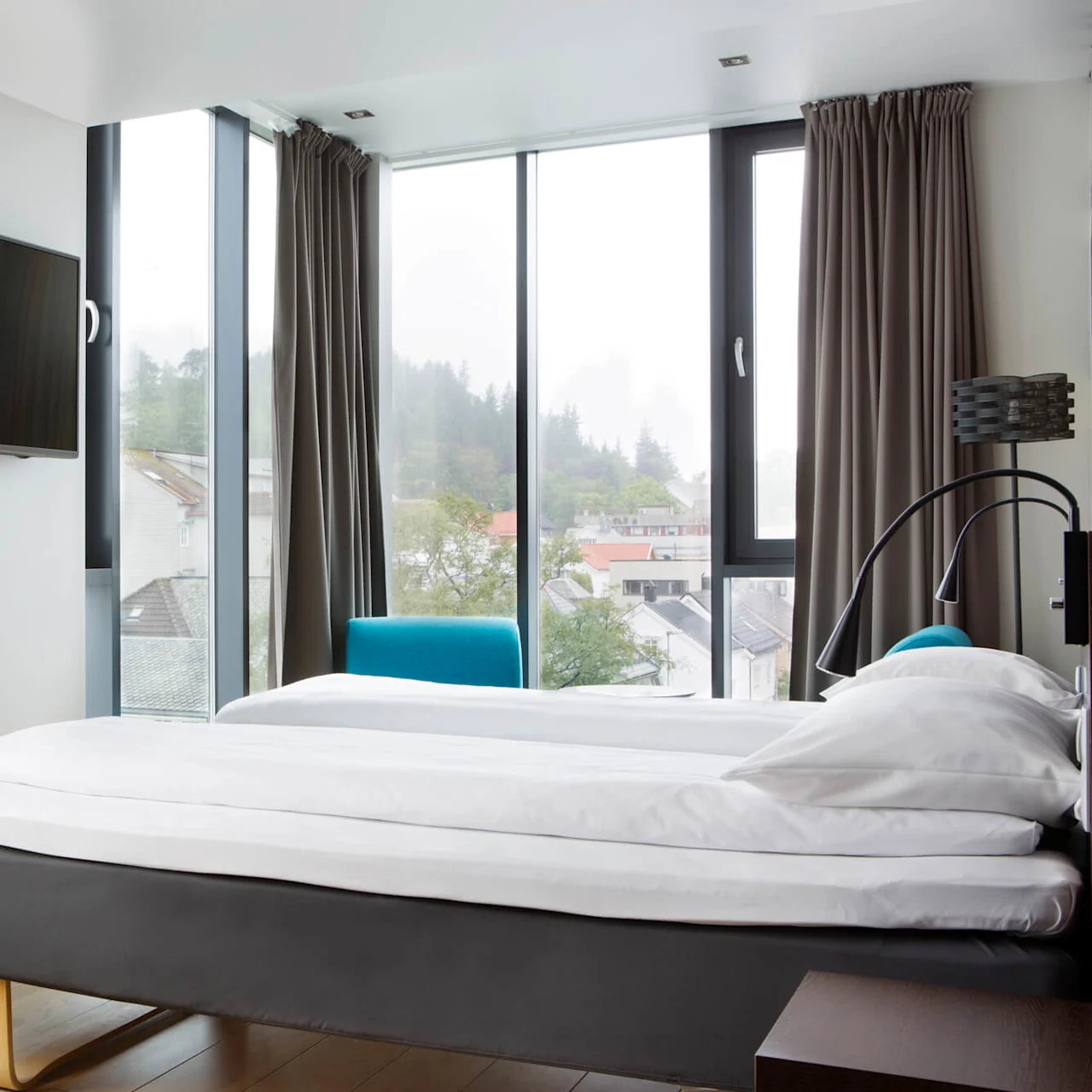 En seng med hvitt sengetøy er plassert mot store vinduer på hotellrommet, med utsikt over en by med trær og bygninger. En veggmontert TV og skrivebord er i nærheten.