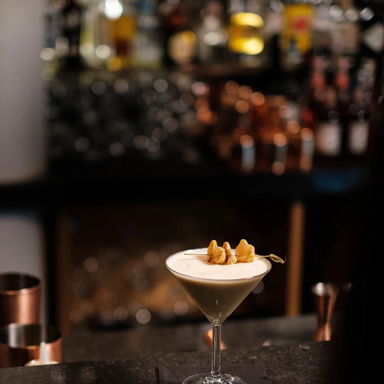A cocktail glass sits on a bar counter, holding a creamy drink garnished with skewered nuts. The dimly lit background shows blurred bottles and copper tools.