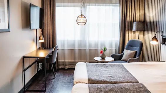 A neatly arranged hotel room features a bed, desk with lamp and chair, armchair with pillow beside a window, and a hanging light. Curtains and striped wallpaper add elegance.