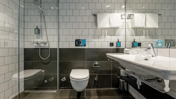 Toalett och handfat i ett modernt badrum med en glasomgiven dusch, vita och grå kakel, handdukar på en hållare och toalettartiklar på bänken, vilket skapar en elegant, ren miljö.