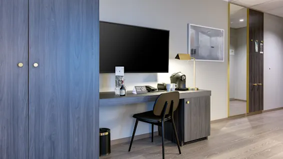 TV på en vegg over et skrivebord med en lampe, kaffemaskin og stol; tre skap ved siden av, moderne hotellrominnstilling. Tekst: "Kitchen," "Thank you for not smoking."
