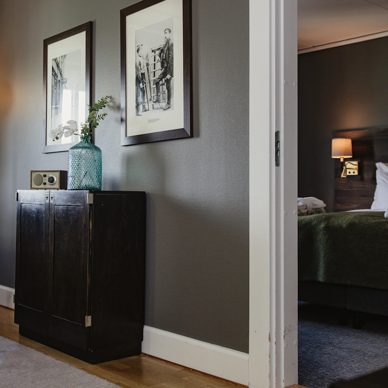 A dark wooden cabinet with a vase and clock sits next to a lit floor lamp; through an open doorway, a bedroom with a neatly made bed and wall lamp is visible.