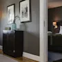 A dark wooden cabinet with a vase and clock sits next to a lit floor lamp; through an open doorway, a bedroom with a neatly made bed and wall lamp is visible.