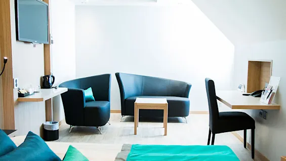 Et moderne hotelværelse har sorte lænestole, en matchende sofa og et sofabord i træ. Et skrivebord er udstyret med en telefon og lampe, mod hvide vægge.
