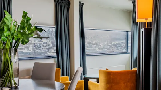 En vase med grønne planter står på et rundt bord. I nærheden er gule lænestole vendt mod store vinduer med byudsigt. Mørke gardiner og en høj lampe forbedrer rummets hyggelige atmosfære.