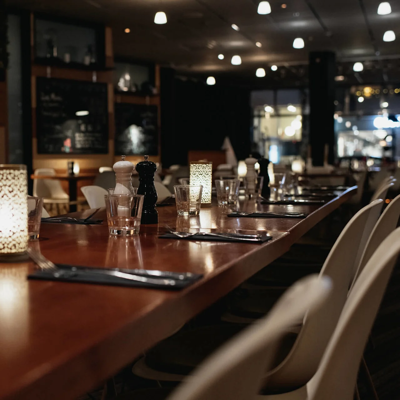 A long wooden table in a dimly lit restaurant is set with glasses, napkins, and candles. Surrounding chairs suggest it's prepared for dining, creating a cozy ambiance.
