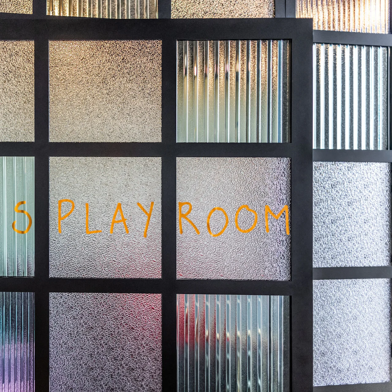 En frostet glassdør med svarte rammer og teksten "Kids Play Room" skrevet med lekne oransje bokstaver, som markerer inngangen til et lekeområde for barn.