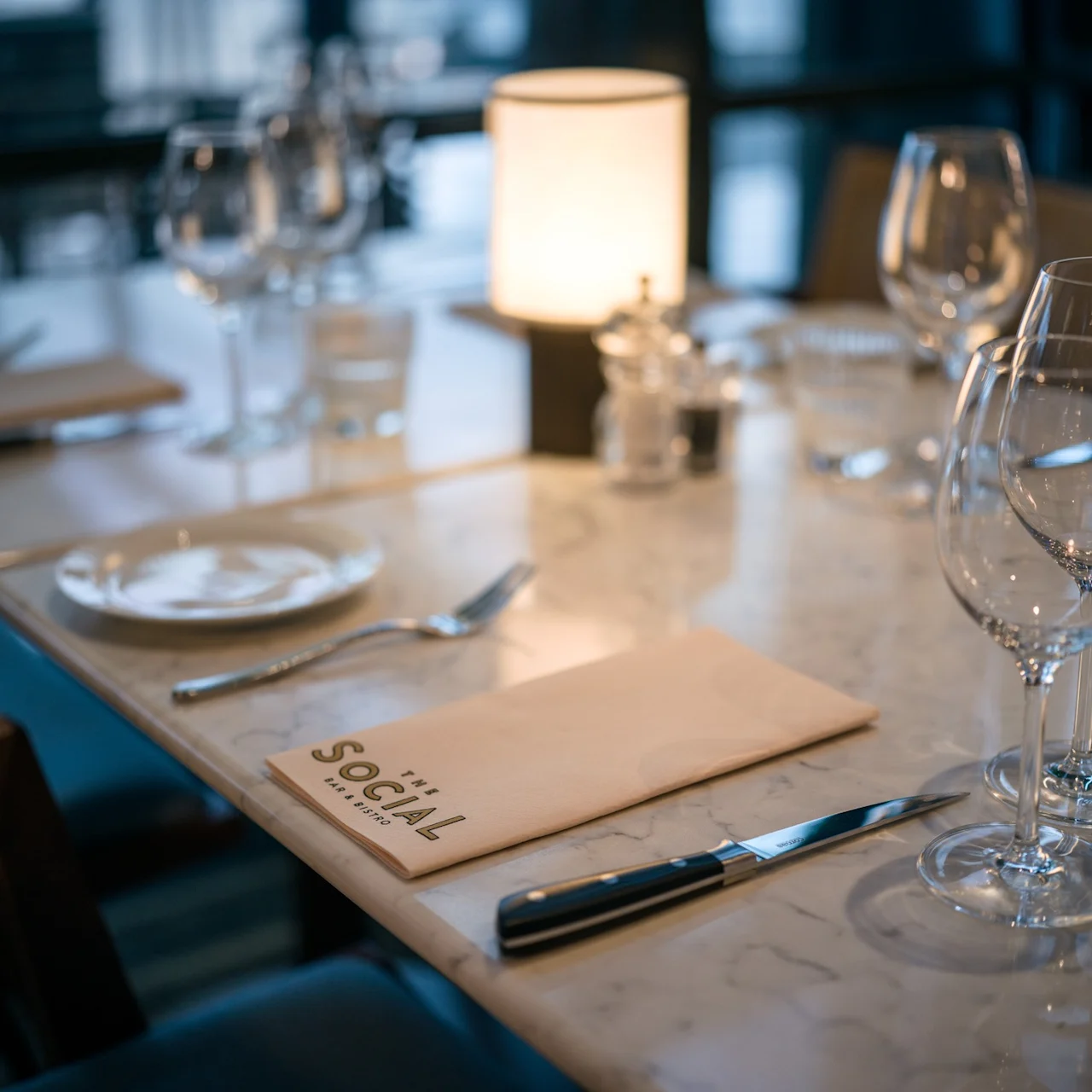 Ett marmorbord dukat med glas, bestick och servetter i en svagt upplyst restaurang. En meny märkt "The Social Wine Bar & Bistro" ligger på bordet.