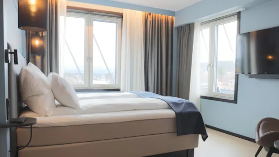 A neatly arranged hotel room with a bed by a window offering a city view, sunlight filtering through curtains, and a wall-mounted TV.