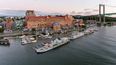 Båtar förtöjda vid en marina bredvid en byggnad med rött tak, omgiven av parkerade bilar. En stor hängbro sträcker sig över vattnet i bakgrunden med grönska längs kusten.