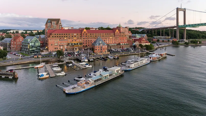 Boats docked on a marina beside a red-roofed building, surrounded by parked cars. A large suspension bridge spans over water in the background, with greenery visible onshore.