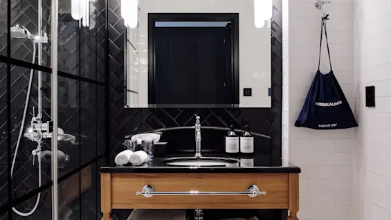 Elegant bathroom with a wooden vanity holding a sink and toiletries. Tiled walls surround with a glass shower visible. Bag labeled "AMERIKALINJEN Hairdryer" hangs nearby. Bright lighting and reflections complete the scene.