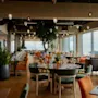 Restaurant med elegant dækkede borde, stole i grøn og orange, og store vinduer, der viser en naturskøn udsigt. Hængende lamper fra loftet forstærker den hyggelige atmosfære. Synlig tekst: "AIME".
