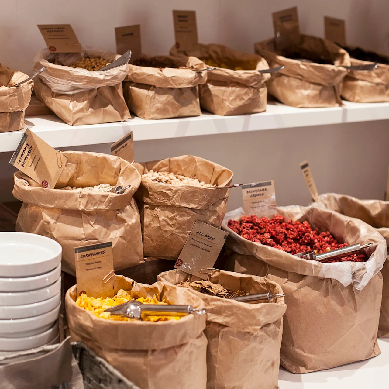 Brune papirposer som inneholder forskjellige kornblandinger med skjeer er pent plassert på hyller i en frokostbuffé. Etiketter lyder: "SOLSIKKE KJERNER," "KORNFLAKES," "ALL BRAN," "MINIFLAKES CRUNCH."