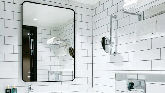 Et rektangulært spejl hænger over en hvid vask i et badeværelse med flisebelagte vægge. Håndklæder hviler på en metalholder, toiletartikler står på vasken, og et toilet er i nærheden.