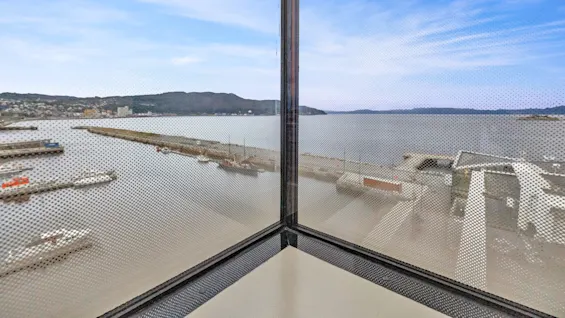 A corner window with dot-patterned glass overlooks a calm harbor with boats docked, surrounded by distant hills under a clear blue sky.