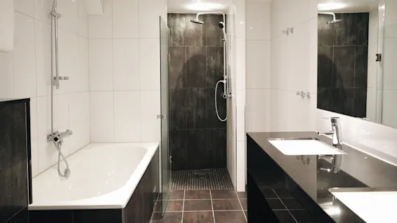 Et moderne bad har et badekar og en glass-omsluttet dusj. En vask med en slank kran står ved siden av et stort speil, omgitt av lyse fliser.