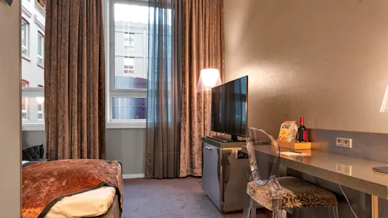 TV’et står på et skrivebord i et moderne hotelværelse med en gennemsigtig stol, brune gardiner og et vindue. En flaske, snacks og lamper giver en hyggelig stemning.