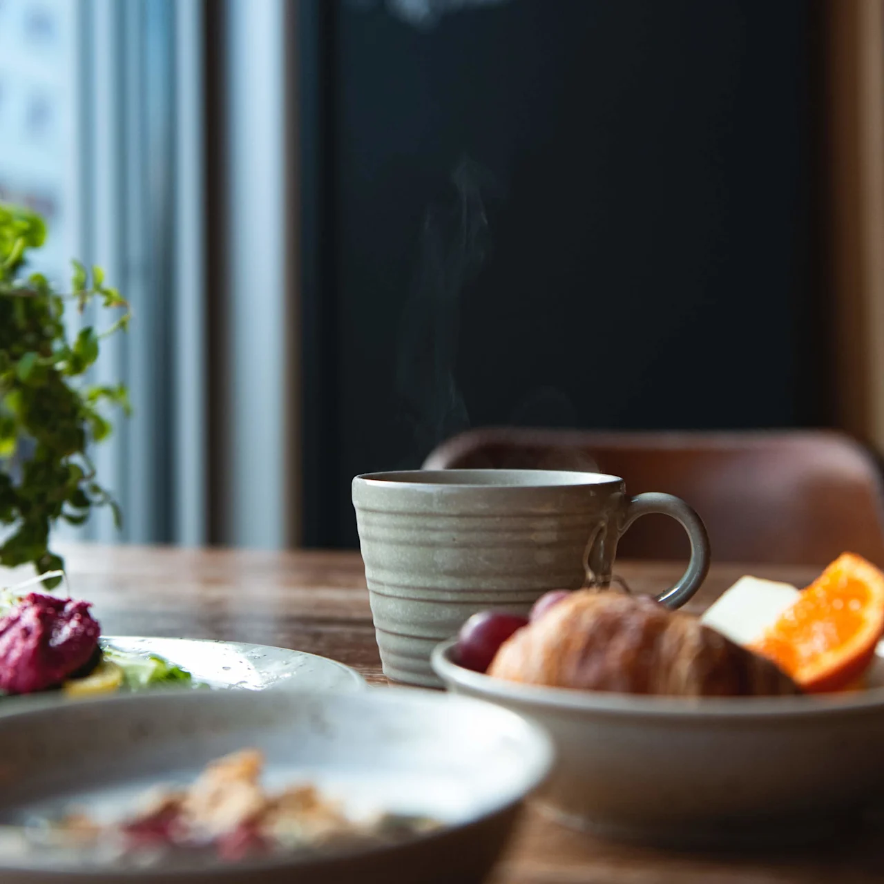 A steaming cup sits on a wooden table, surrounded by a croissant, fruit, and greenery. In the background, a chalkboard reads "SPECIALS of the MONTH".