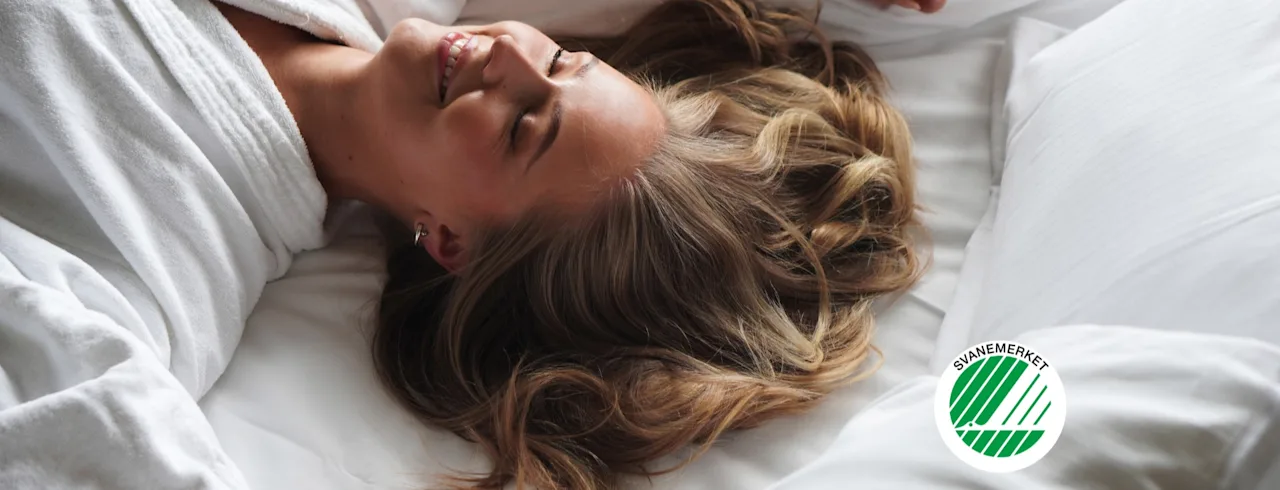 A person happily relaxing in a white bathrobe on a bed with a Svanemerket pillow.