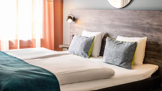 A neatly made bed with two gray and white pillows sits in a cozy bedroom, illuminated by a small wall lamp, near sheer pink curtains allowing soft light.