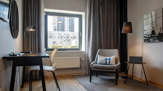 A desk with a black lamp and chair sits by a window in a modern room. Nearby, an armchair with a pillow and a side table enhance the cozy atmosphere.