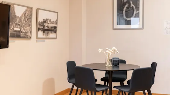 Et koselig hjørne med et rundt bord, fire stoler og innrammet kunst på veggene, med en vase med tørkede blomster.