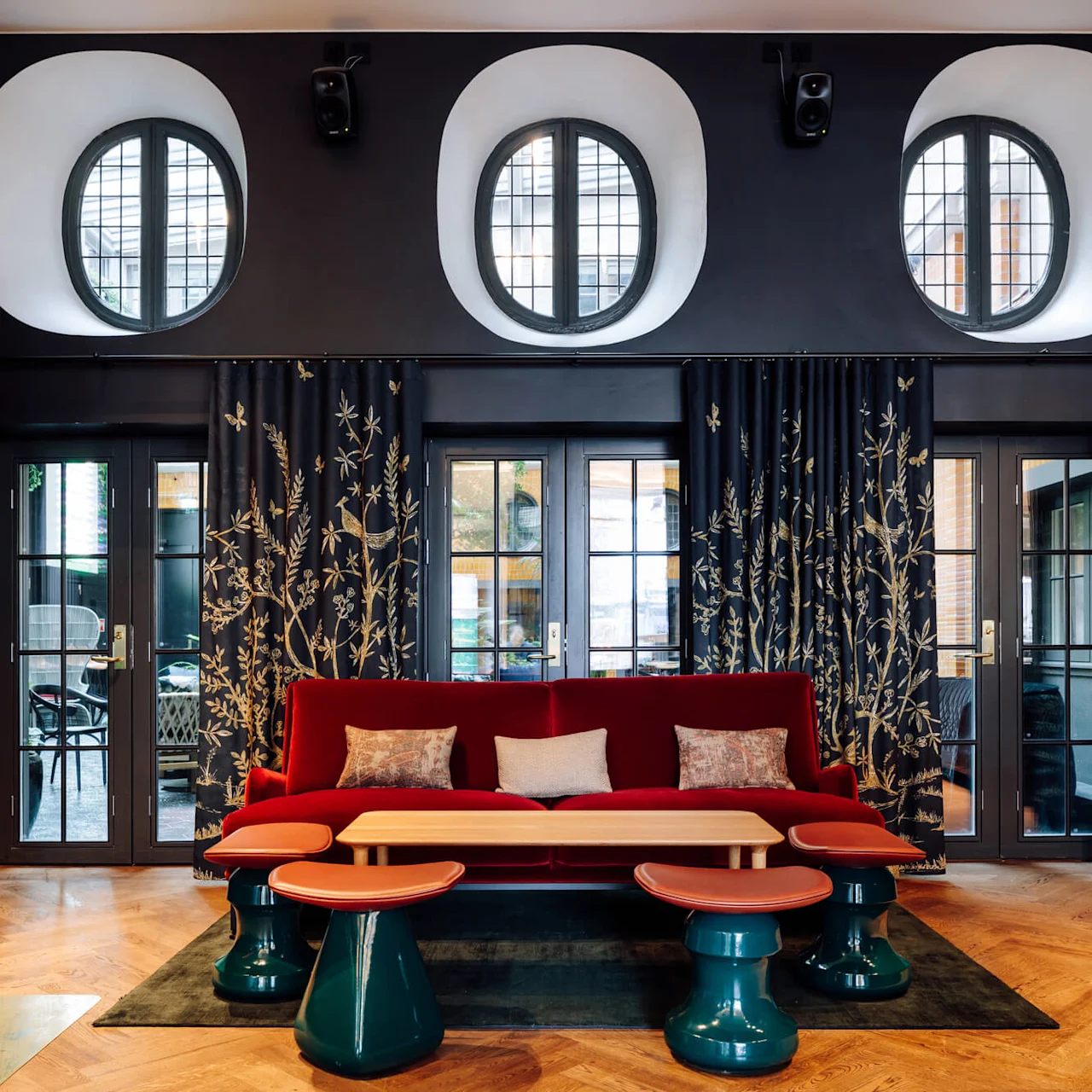 Red sofa with cushions against a wall of circular windows. Ornate curtains feature floral patterns. Surrounding are glass doors and teal, round tables. A modern fireplace is on the left.