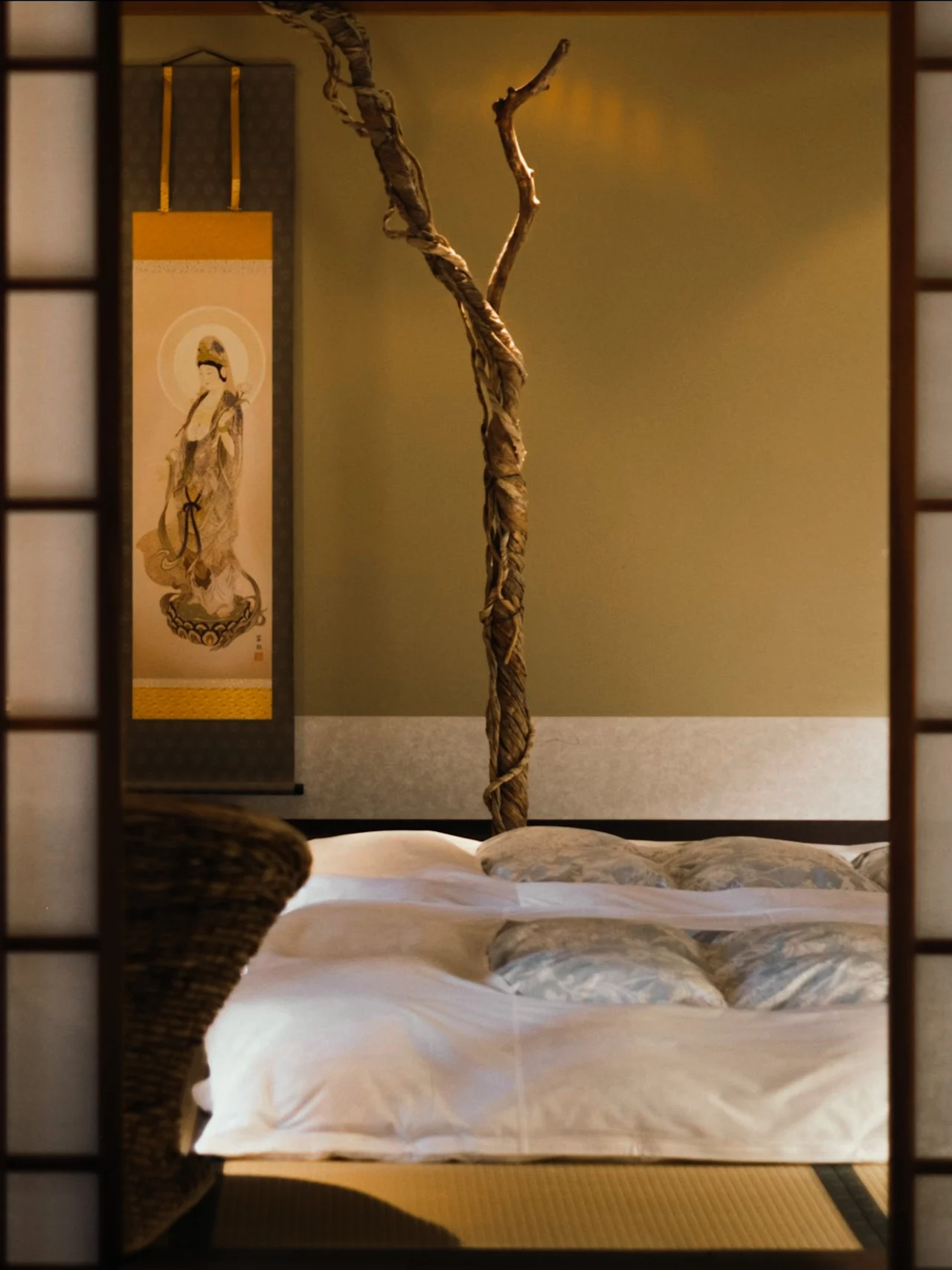 Rauhallinen makuuhuone shoji-näytön läpi, jossa on pehmeä sänky, koristeellinen oksa ja perinteinen rullamaalaus.