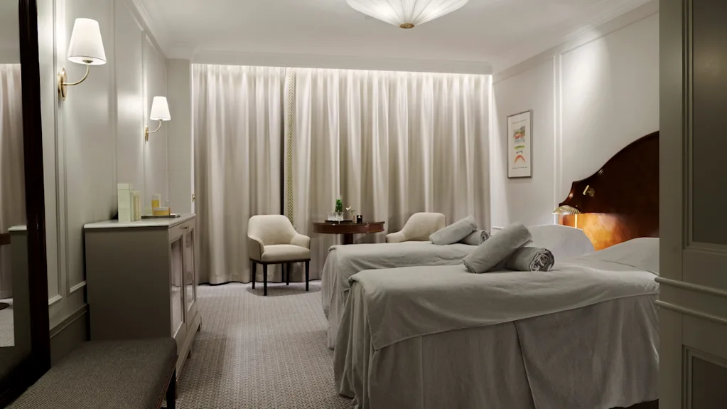 Ett hotellrum har två prydligt bäddade sängar med grå sängkläder, sänglampor som lyser, en sittgrupp med stolar och bord nära ett fönster med fördragna gardiner.