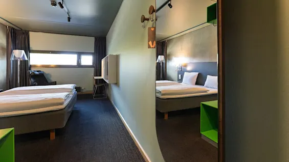 A hotel room features a double bed with white linens; a chair and desk are visible near a window. A wall mirror reflects the bed and green shelves. 