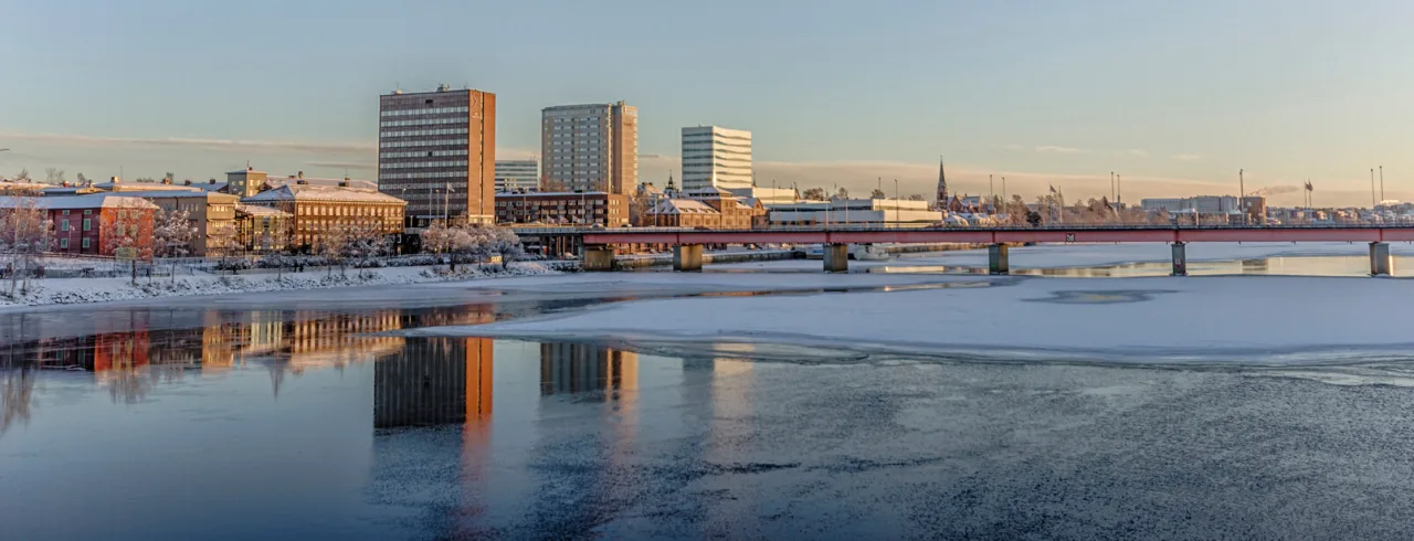 En bysilhuet med bygninger, der reflekteres på delvist frossent flodvand under en klar blå himmel ved daggry eller skumring.