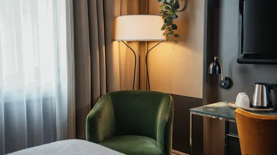 Et hyggeligt hotelværelseshjørne med en grøn lænestol, en varm lampe og et skrivebord med en elkedel, der indbyder til afslapning.