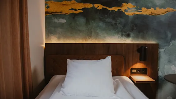 En komfortabel hotellseng med en hvit pute, sengegavl i tre, og en vegg med abstrakt kunst i gull- og grønntoner.