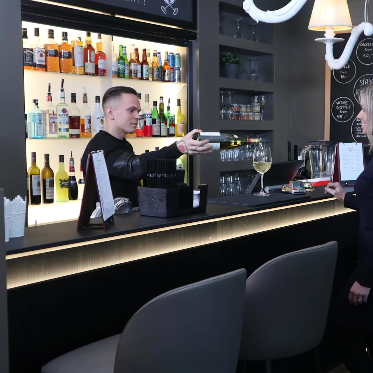 A bartender is handing a card to a customer at a modern bar with shelves of bottles and a blackboard listing drinks.