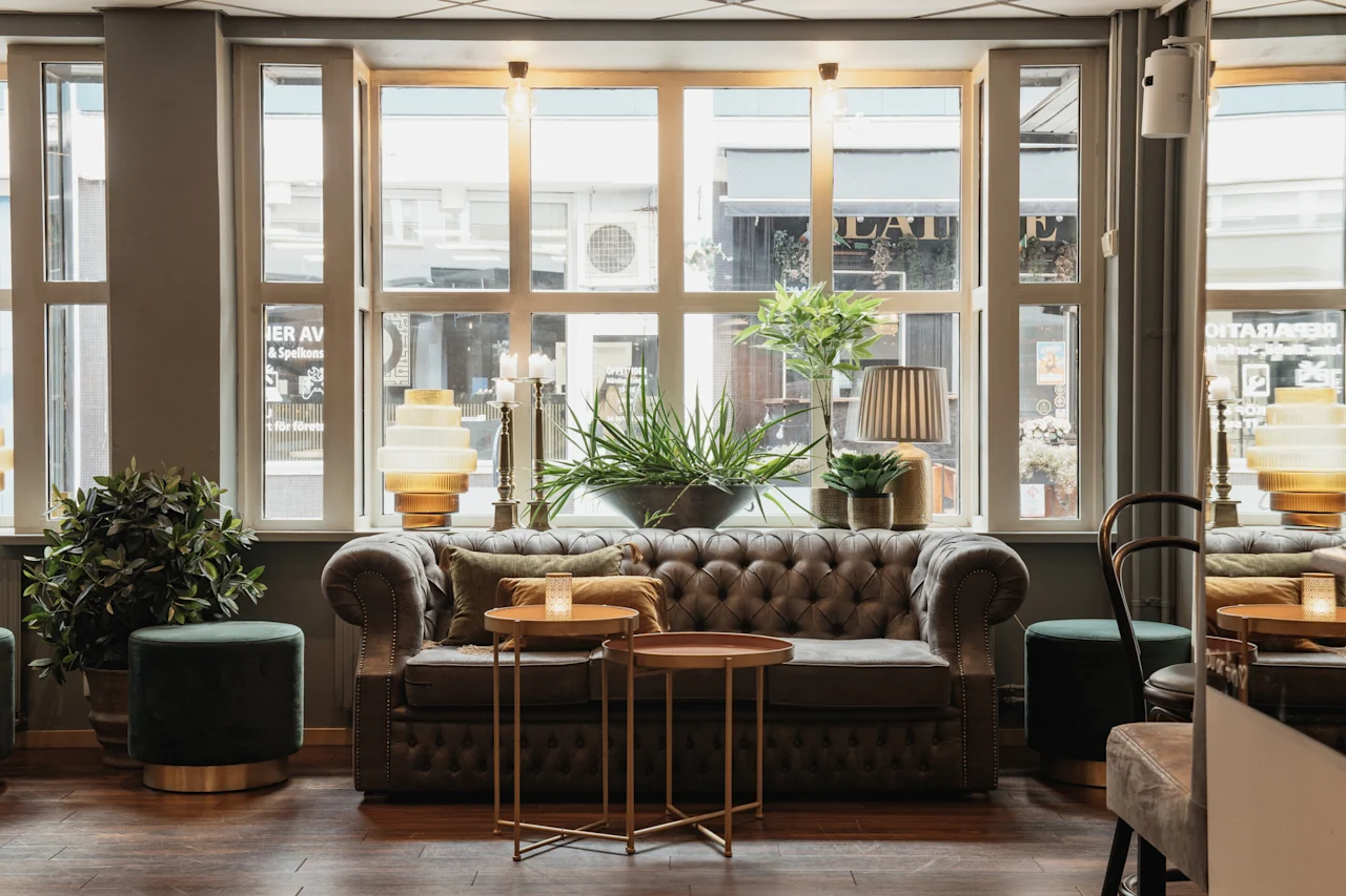 En bekväm lounge med en tuftad soffa, växter och varm belysning vid ett fönster. Text synlig genom fönstret: IER AV Spalkons, LAI, OITARABAS.