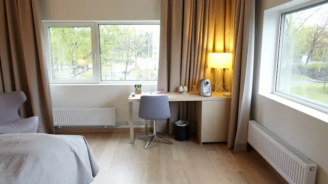 Et skrivebord står i et lyst rom, opplyst av en lampe. Det har en kaffemaskin, kopp og bilderamme ved siden av en stol. Store vinduer viser grønt utenfor.