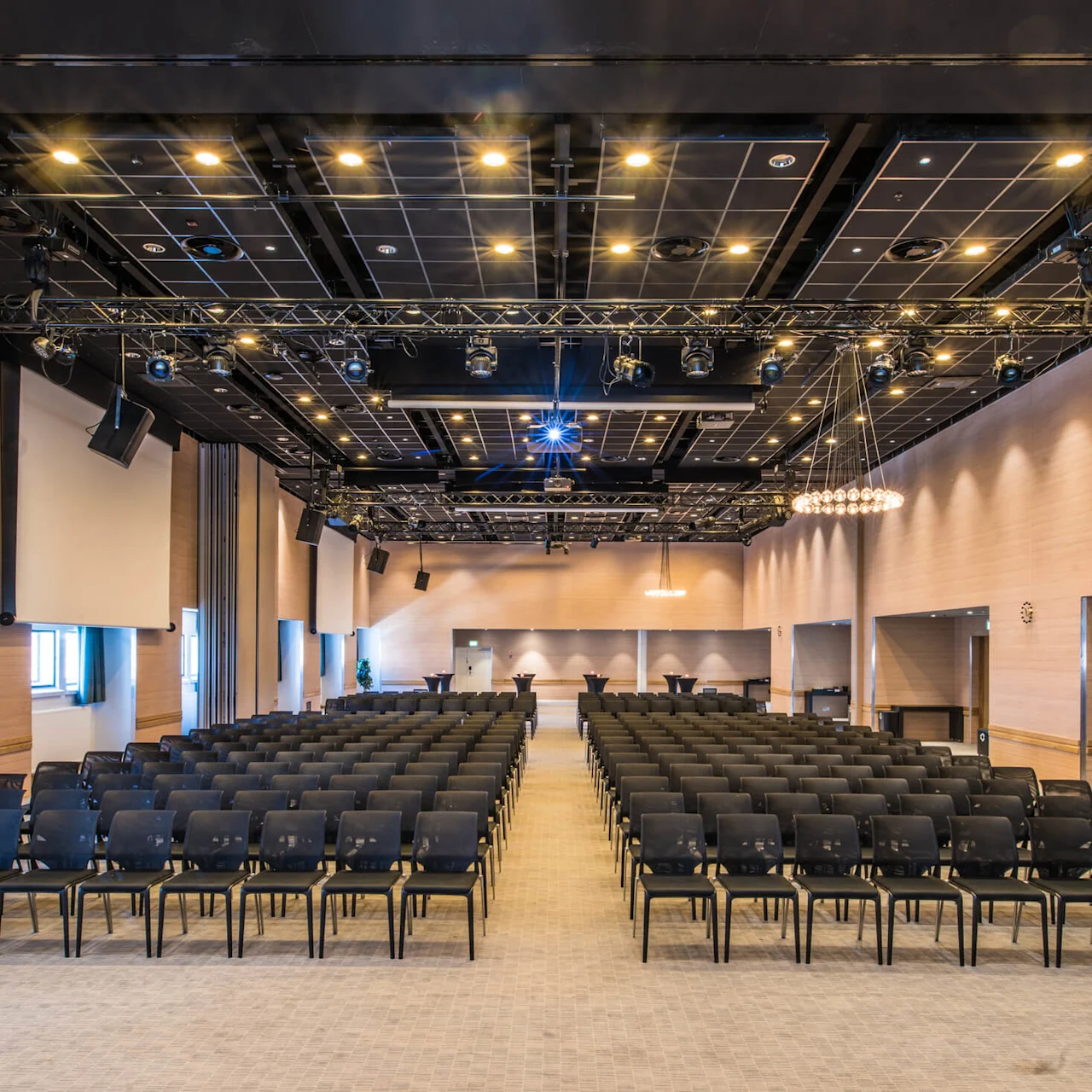 Et stort konferencelokale med rækker af sorte stole vendt mod en scene. Loftet har adskillige spotlights og hængende udstyr. Væggene er af træ, hvilket tilføjer varme til det rummelige miljø.